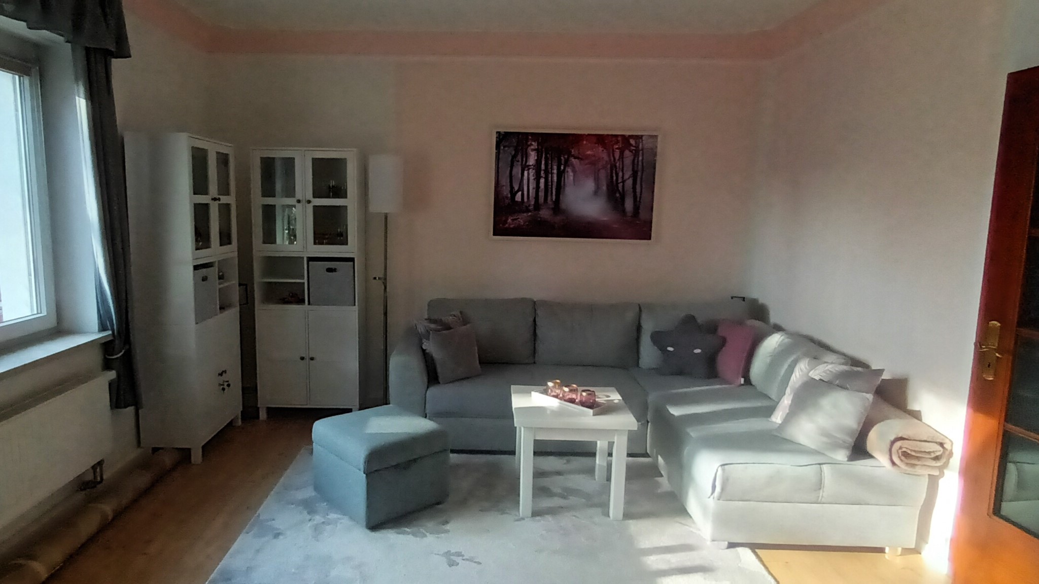 Gemütliches Wohnzimmer mit grauer Couch, weißen Möbeln und Wald-Wandbild. So funktioniert modernes Wohnen.